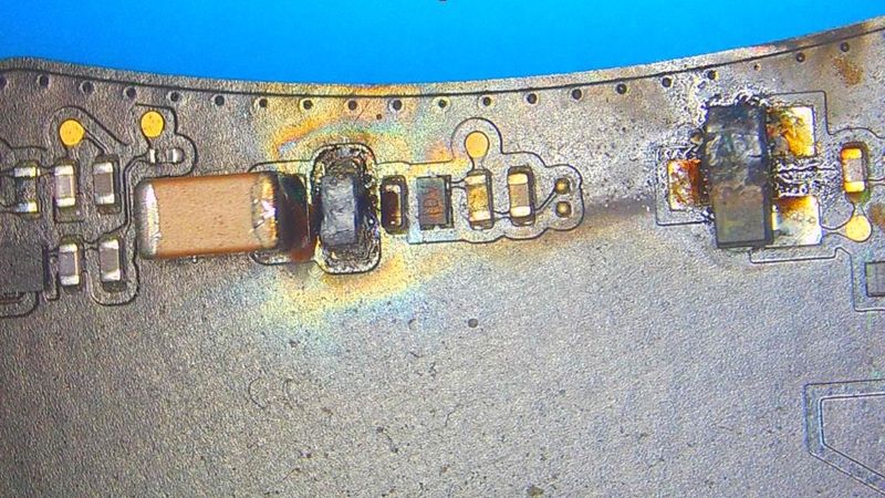 Vista al microscopio de una placa electrónica durante microsoldadura, con componentes SMD resoldados y marcas de calor alrededor