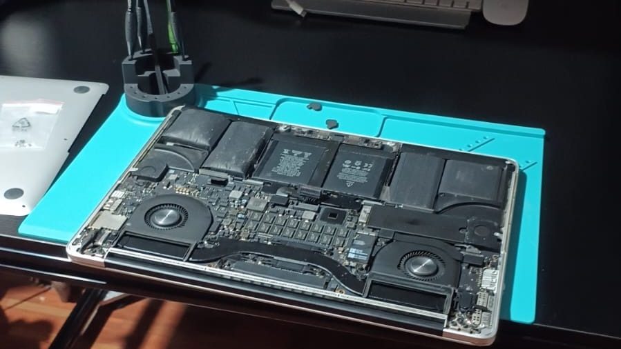 MacBook Pro abierta sobre un tapete antiestático en el taller, con ventiladores, baterías y placa lógica a la vista para mantenimiento
